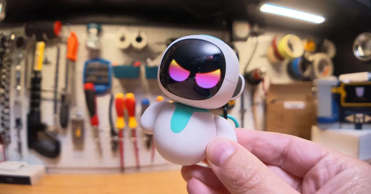 A cute little robot toy with scary rainbow eyes, held with my thumb and forefinger, with my garage workbench in the background.